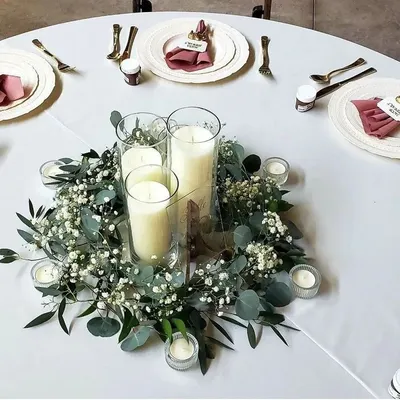 A classic and elegant table centerpiece. The arrangement features white pillar candles in glass vases, surrounded by a wreath of baby's breath and eucalyptus leaves.