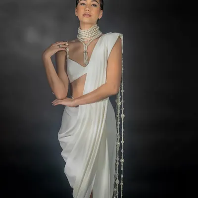 A studio shot for Nikhil Thampi's 'Gleam Collection', focusing on a modern white saree. The lighting is sculpted to highlight the intricate pearl jewelry and the unique drape of the garment against a dark, moody backdrop.