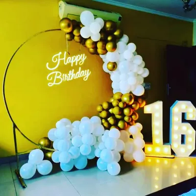 A chic 16th birthday setup with a gold and white balloon arch on a ring frame. The large light-up '16' numbers and 'Happy Birthday' neon sign make it a perfect spot for birthday photos.