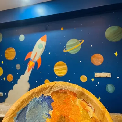 A work-in-progress shot of the solar system mural. You can see my paint palette in the foreground, filled with the cosmic colors used to bring the planets and rocket ship to life on the deep blue wall.