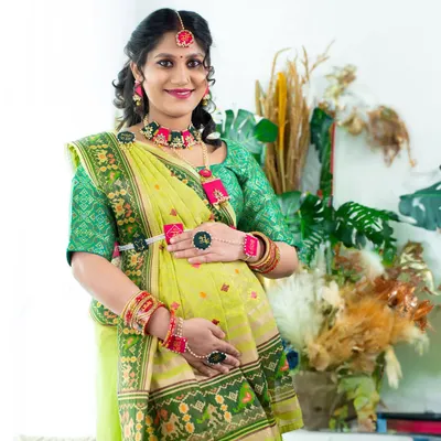 A vibrant and traditional portrait. The mom-to-be is dressed in a beautiful green saree with intricate details and matching floral jewelry, celebrating her pregnancy with cultural pride.