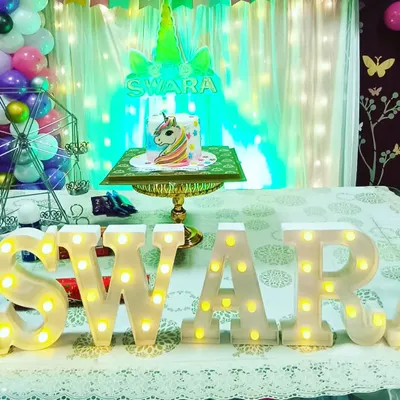 A magical unicorn theme for Swara's birthday. The setup features a beautiful cake table with a unicorn cake, marquee letters of her name, and a backdrop with fairy lights, creating a whimsical photo corner.