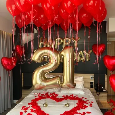 A classic romantic birthday surprise featuring a bold red and gold color scheme. The decoration includes large "21" foil balloons, "Happy Birthday" lettering, and a heart made of rose petals on the bed.