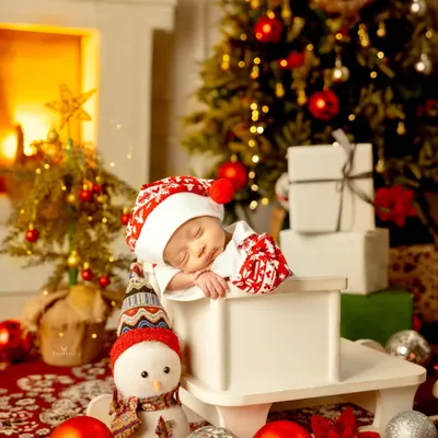 A perfect Christmas dream. This newborn sleeps peacefully in a festive setup with a warm fireplace, a decorated tree, and shiny ornaments.