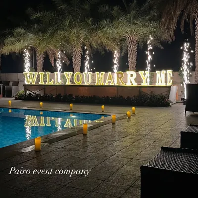 A stunning poolside proposal at night, featuring large illuminated "WILL YOU MARRY ME" letters, a path of electronic candles, and celebratory cold pyro fireworks in the background.
