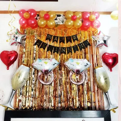 This engagement party decoration is perfect for celebrating a new beginning. The setup includes a gold tinsel curtain, foil balloons shaped like diamond rings and champagne glasses, and a celebratory banner.