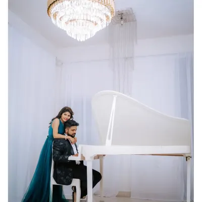 An elegant pre-wedding portrait of a couple in western attire, posing by a grand white piano under a magnificent crystal chandelier.
