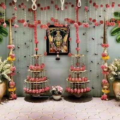An elegant decor setup for a bridal pooja at home. The design uses traditional brass lamps (deepams), strings of jasmine and pink flowers, and a framed picture of a deity to create a serene and sacred space.