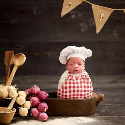 Introducing the world's tiniest chef. This newborn is dressed in a chef's hat and apron, sitting in a cooking pot surrounded by wooden utensils and vegetables.