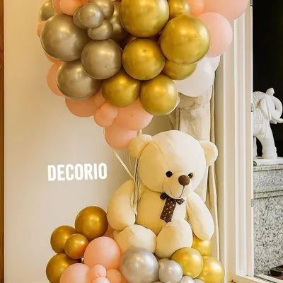 A simple yet beautiful balloon arrangement to welcome a new baby. A large teddy bear is seated within a cluster of pink, gold, and silver balloons, perfect for placing at the entrance of your home.