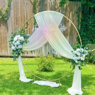 A romantic outdoor backdrop perfect for an engagement or daytime wedding ceremony. The golden ring frame is elegantly draped with sheer fabric and adorned with white floral arrangements, with a "Happily Ever After" neon sign.