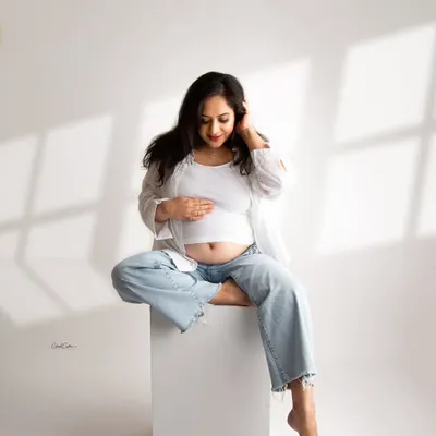 A serene and modern maternity portrait. The mom-to-be is dressed in a simple white top and jeans, sitting peacefully in a brightly lit studio setting.