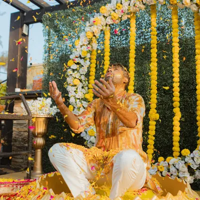 A groom raises his hands in celebration as he is showered with turmeric and flower petals. This action shot perfectly captures the energy of a Haldi ceremony.