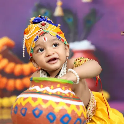 A close-up of a baby dressed as Little Krishna, playfully looking out from behind a butter pot. The detailed costume and traditional props make this a truly divine portrait.