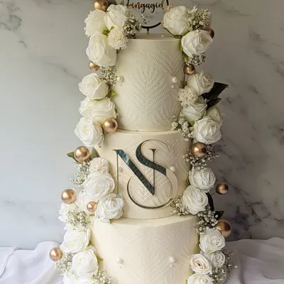 A grand three-tier engagement cake designed for a lifetime of memories. The design features a cascade of fresh white roses and baby's breath, a custom monogram, and delicate stenciling for a look of pure sophistication.