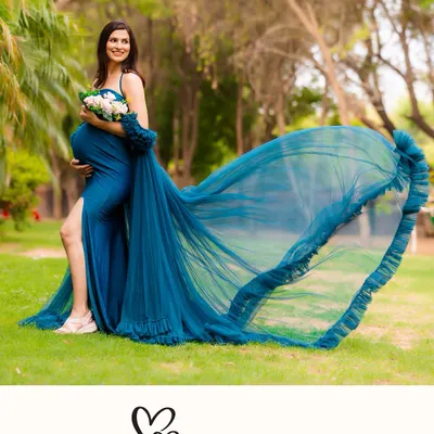 A stunning outdoor shot of a mother-to-be in a vibrant blue gown with a dramatic, flowing train. Holding a bouquet, she smiles, surrounded by the lush greenery of a park.