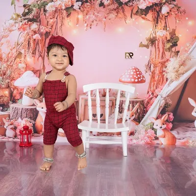 A baby boy stands proudly in a whimsical woodland setup, complete with a teepee, mushrooms, and friendly animal figures. The soft pink and red tones create a warm and magical forest scene.