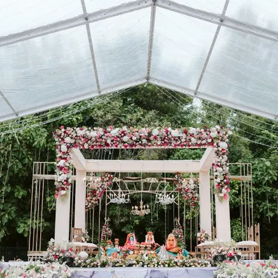 A breathtaking mandap setup inside a clear marquee tent, surrounded by lush greenery. The modern white structure was adorned with beautiful red and white florals and crystal chandeliers for a truly dreamy wedding ceremony.