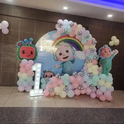 A cheerful Cocomelon theme for a first birthday. This setup features a custom backdrop with a rainbow and Cocomelon characters, a pastel balloon arch, and a large light-up number '1'.