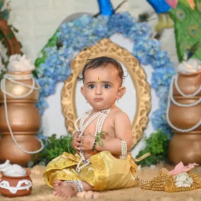 A classic Little Krishna pose. This setup features traditional pots, peacock feathers, and a beautiful arch, creating a perfect backdrop for this baby dressed as Krishna, complete with pearl necklaces.