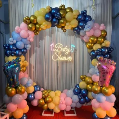 A vibrant gender reveal setup with a circular balloon arch in pink, blue, yellow, and gold. A "Baby Shower" neon sign and "It's a Boy/Girl" foil balloons add to the excitement.