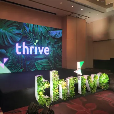 An angled view of the 'thrive' corporate stage. The large LED screen displays branded content, while the custom 3D greenery-filled letters provide a unique, on-theme focal point that is both professional and visually interesting.