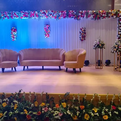 This is a modern and chic stage design for a sangeet or reception. It features stylish pastel pink velvet sofas against a clean white curtain backdrop, accented with colorful floral arrangements and soft blue lighting.