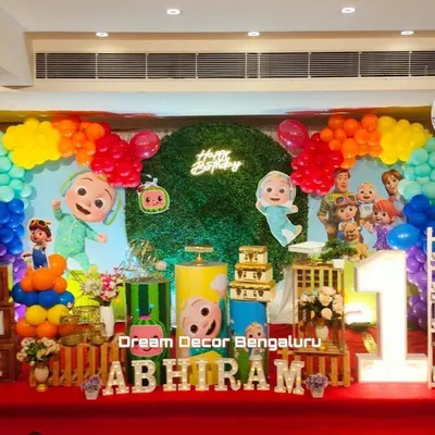 The bright and cheerful world of Cocomelon is perfect for a first birthday party. This setup features a lush green wall, a rainbow balloon garland, and all the beloved characters, including the iconic yellow school bus.
