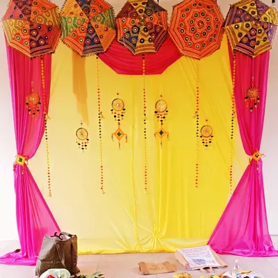 A classic Haldi decor setup with a bright yellow backdrop, pink drapes, and colorful Rajasthani umbrellas. The hanging ornaments add a festive, traditional touch.