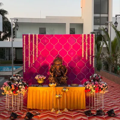 A grand welcome setup for Lord Ganesha, featuring a vibrant pink backdrop with a traditional pattern. The idol is placed on a yellow-draped table, flanked by large floral bouquets and spotlights.