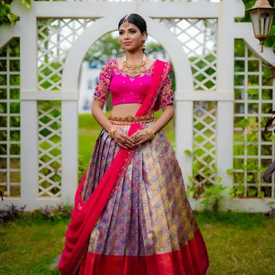 A full-length shot showcasing a vibrant Kanjeevaram half saree. The outdoor garden setting provides a beautiful, natural backdrop that complements the bright colors of the bridal lehenga, ideal for a designer catalog.