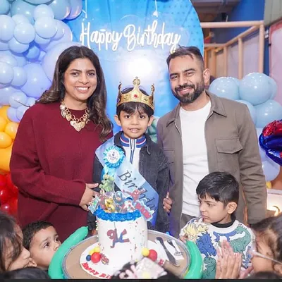 The birthday boy, wearing his crown and sash, stands with his proud parents for the cake-cutting ceremony at his superhero-themed party.