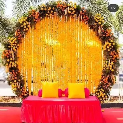 A classic and grand Haldi backdrop featuring a large circular frame completely covered in marigold flower strings. The bright yellow and orange tones create a traditional and cheerful setting for the ceremony.