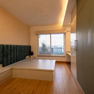This bedroom is designed for tranquility, with a plush green velvet headboard and a platform bed that includes built-in side tables. The large window with blinds allows for adjustable natural light, creating a calm and restful ambiance.