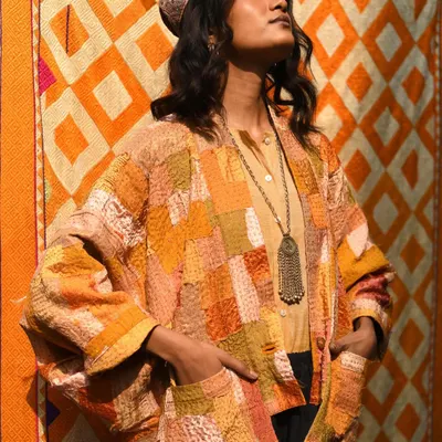 Bathed in warm light against a vibrant, patterned backdrop. This portrait for Wandering Silk captures the rich, earthy tones of a patchwork Kantha jacket, highlighting its texture and artisanal feel.