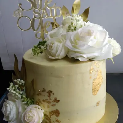 A classic engagement cake for a "She Said Yes" moment. This design features a soft yellow buttercream finish, delicate gold leaf accents, and is topped with beautiful, large white silk roses for an elegant and celebratory look.
