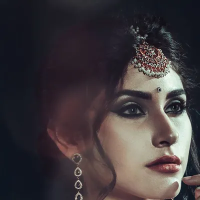 A dramatic close-up where the traditional maang tikka is the hero. The dark, moody lighting is intentional, designed to make the intricate details of the jewelry and the model's smokey eye makeup stand out.
