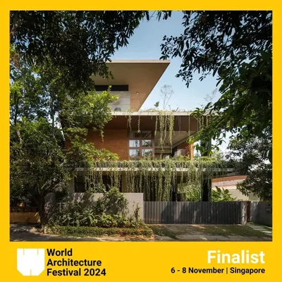 The 'House of Greens' seen from the street, framed by trees. The design uses a combination of exposed brick, concrete, and abundant greenery to create a structure that feels rooted in its natural surroundings.