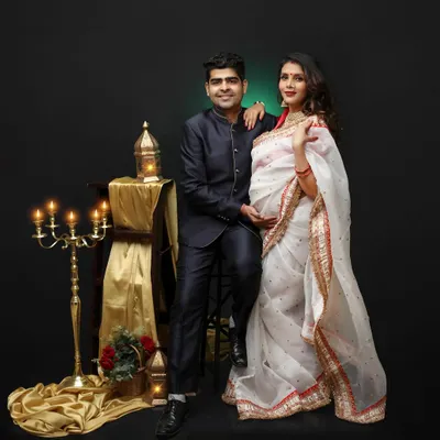 A beautiful couple's portrait celebrating their Indian heritage. The mom-to-be looks stunning in a white and red saree, complemented by traditional props and warm, dramatic lighting.