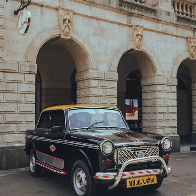 The timeless charm of a classic "kaali-peeli" Premier Padmini taxi, parked in front of the beautiful arches of a heritage building in Mumbai.
