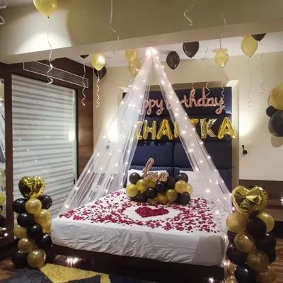 A complete romantic room setup for a birthday. This decoration features a bed canopy with fairy lights, a heart shape made of rose petals on the bed, and black and gold balloon arrangements.