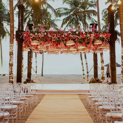 A stunning beach mandap setup facing the ocean. The structure is heavily adorned with pink and red florals, with traditional umbrellas hanging down, creating a perfect frame for a sunset ceremony.