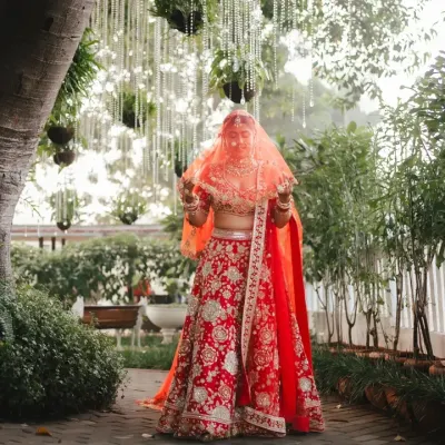A bride veiled in red, standing amidst a lush green garden with crystal hangings. This portrait is the epitome of bridal grace, a mysterious and beautiful moment before she walks down the aisle.