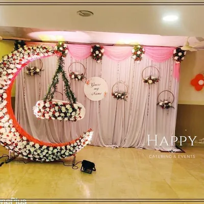 A beautiful crescent moon cradle decorated with white and red flowers, set against a soft pink and white draped backdrop for a naming ceremony. A "Guess my name" sign adds a playful touch.