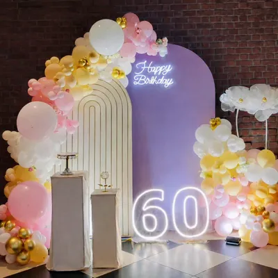 Celebrating a 60th birthday in style with this sophisticated setup. It features a pastel balloon arch, a modern arched backdrop, and large, illuminated marquee numbers, all tied together with a "Happy Birthday" neon sign.