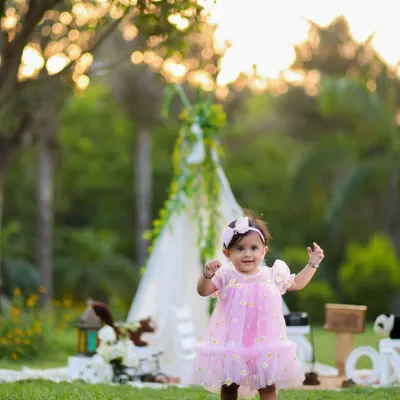A beautiful outdoor portrait of a one-year-old girl in a pink dress, taking her first steps in a lush green park. The teepee in the background adds a touch of whimsy.