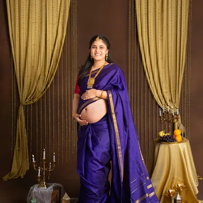 A beautiful traditional maternity portrait. The mother-to-be is dressed in a royal blue silk saree, her baby bump peeking out, in a studio setting with classic golden drapes and brass decor.