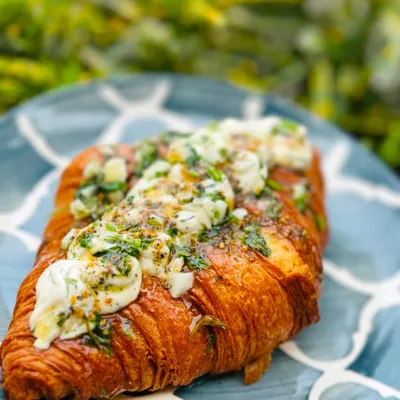 A close-up of our Korean Cream Cheese Croissant. This savory treat is filled with a slightly sweet cream cheese and topped with a delicious garlic herb butter, a must-try from our menu.