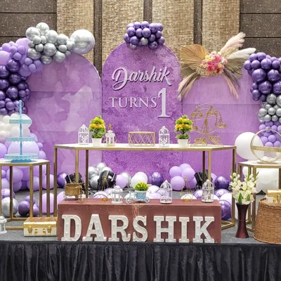A beautiful first birthday stage in a purple and silver color scheme. The setup includes a custom backdrop, balloon arches, a dessert table, and light-up letters for the child's name.