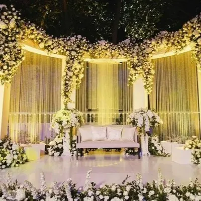 An enchanting outdoor reception stage completely enveloped in white flowers. The cascading floral arrangements and soft, warm lighting create a dreamy and romantic atmosphere under the night sky.